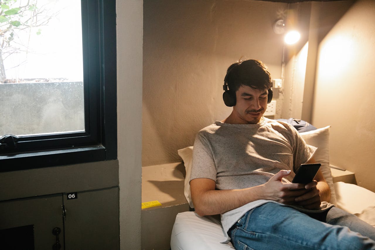 Young male traveller resting on comfortable bed and listening to music in headphones while browsing mobile phone during trip in train compartment