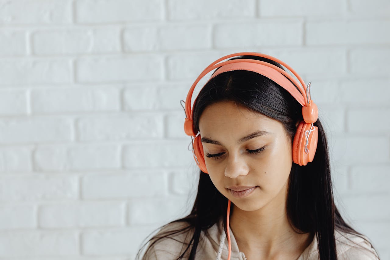 Portrait of a serene young woman wearing orange headphones against a textured white brick wall, eyes closed.